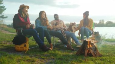 Göl kenarındaki kamp ateşinde çeşitli arkadaşlar altın saat boyunca dinleniyor. Kot giyen beyaz kadın, hasır şapka takan beyaz adam.,