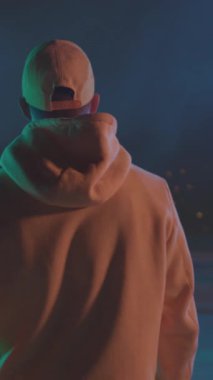 Geceleri çatıdaki siluet, sarı kapüşonlu beyaz erkek hareketsiz duruyor ve uzak şehir bokeh, deniz mavisi kenarını izliyor.