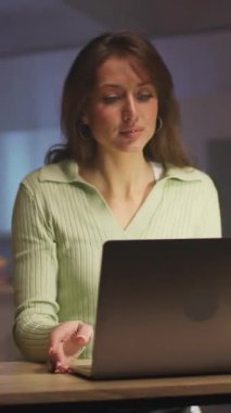 Laptop 'ta çalışan beyaz bir kadın, yumuşak lamba parıltısı ve konsantre ifade, rahat giyinirken klavyeye notlar yazma