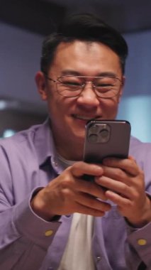 Telefona gülümseyen Asyalı adam, iç mekan aydınlatması, mor gömlek ve gözlük, videodaki kahkahaları yakalıyor.