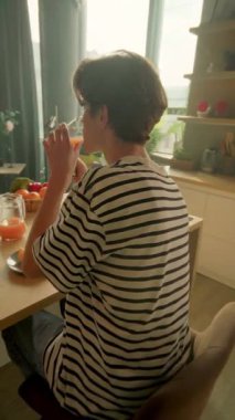 Telefon ve meyveli güneşli mutfak barı. Mesajlaşırken ve kaydırırken gülümseyen genç adam.,