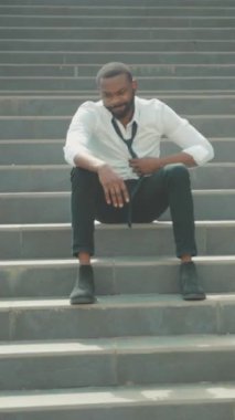 Düşünceli siyah adam basamaklarda oturuyor, başka tarafa bakıyor, kravatını omzuna takmış, beyaz gömlek, yakınlarda küçük kutular, planlar yapıyor.