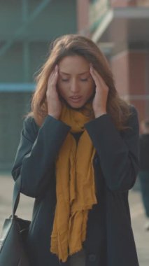 Ofis cephesinde alnını tutan beyaz bir kadın, hardal atkısı ve koyu bir ceket giyiyor, yakından bakınca endişeli bakışlar gösteriyor.