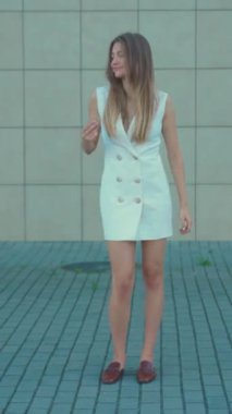 Beyaz kolsuz elbise giymiş, şakacı hareketlerle süslü duvara poz veren genç bir kadın.