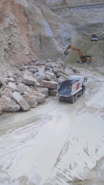 Kazıcı büyük kayaları kamyona yüklüyor, hidrolik kol kürekle düzensiz kayaları topluyor ve kargo yatağına yerleştiriyor.