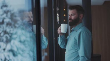 Pencere kenarında duran beyaz adam, kahve sakallı girişimci karla kaplı ağaçlara bakarken sıcak bir bardak tutuyor.