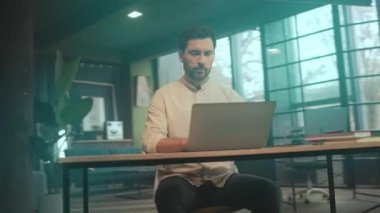 White man focused at laptop desk, natural light, large windows, indoor plants, muted teal grading, typing on keyboard,