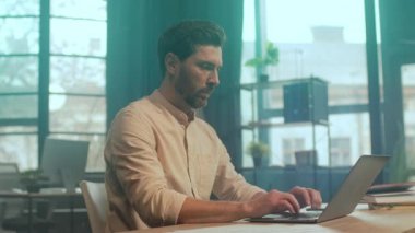 Beyaz adam pencerenin yanında yazmaya odaklanmış, şehir manzarası camdan, güneş ışığından, hesap tablosu olan laptop ekranından.,