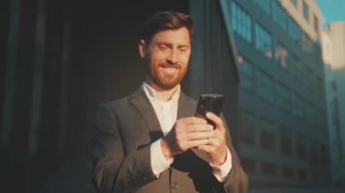 Şehirdeki beyaz girişimci akıllı telefondan kutlama yapıyor. Altın saat arkadan ışıklandırılmış portre, özel dikim takım elbise ve sakal.