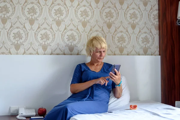 Relaxed woman is sitting on a bed in a hotel holding a smartphone in her hand. Online purchases to check your mobile app or social network. Selective focus