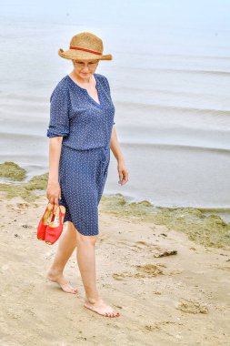 Hasır şapkalı mutlu yetişkin kadın deniz kenarında yürüyor..