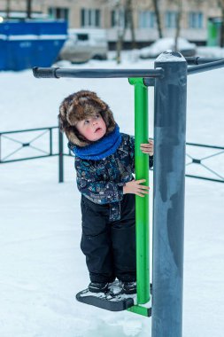Tulumlu 3 yaşında bir çocuk yetişkinler için açık hava egzersiz makinesinin üzerinde duruyor. Kış yürüyüşü.
