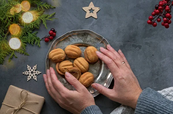 Women\'s elegant hands touch a plate of cookies-nuts on a dark table. Framed layout of Christmas decorations, gifts, nuts and thuja tree. Christmas layout