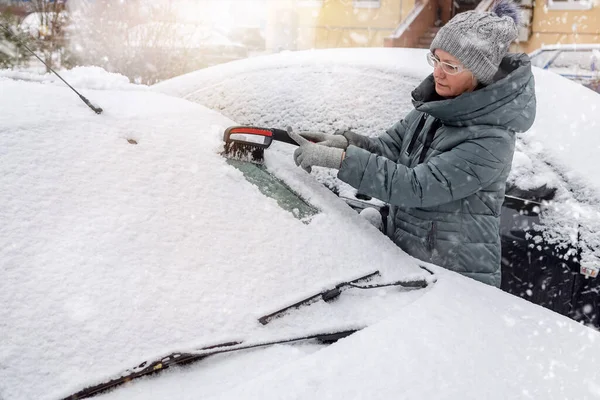 woman cleans snow from the windshield of a car outdoors on a winter day.