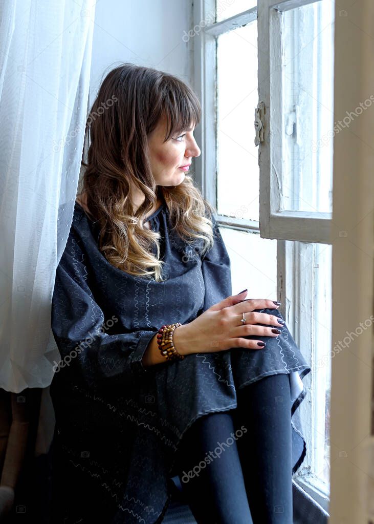 young brunette woman in a dress sits dreamily by the window.