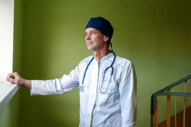 Beyaz üniformalı bir erkek doktor hastanenin merdivenlerinde duruyor..