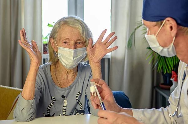 A caring doctor helps a negative elderly grandmother of 85 years-a patient at home or in the hospital. The concept of health care for the elderly. Denial of vaccination
