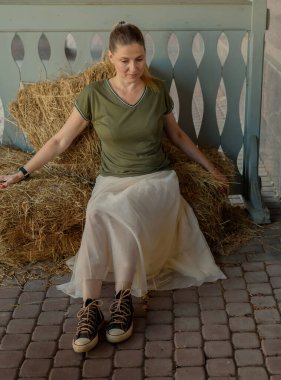 Uzun saçlı, uzun etekli, tişörtlü genç bir kadın yazın ekolojik bir kafede samanların üzerinde oturuyor. Kırsal kesimde bir kadının portresi. Ekolojik eko-turizm kavramı