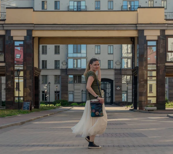 一个穿着长裙 提着袋子穿过街道的年轻漂亮的女人 望着别处 夏天在圣彼得堡散步的女孩 — 图库照片