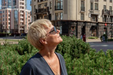 İş, tüketim ve insan kavramı - modern evlerin arka planına karşı güneş gözlüğü takmış yaşlı bir kadın. Büyük Şehir Emlakçısı.