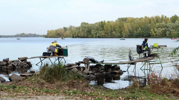 A fisherman catches fish on the river, rear view, from the bank. A fisherman sits on a wooden and stone bridge on the river bank and tries to catch fish. Sports, recreation, lifestyle.