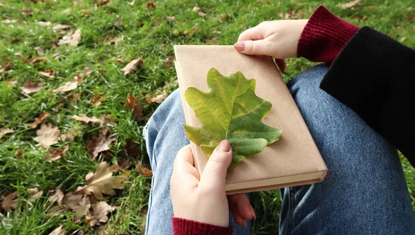 A woman holds a closed book lying in her lap with a fallen oak leaf close-up in a park on a sunny warm autumn day. The concept of relaxation, reading and relaxation alone