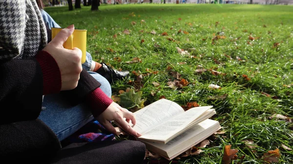 A girl sits on a blanket in the autumn forest, reads a book and drinks a hot drink from a yellow cup in a city park on a warm day on a green meadow. The concept of reading, relaxation and comfort