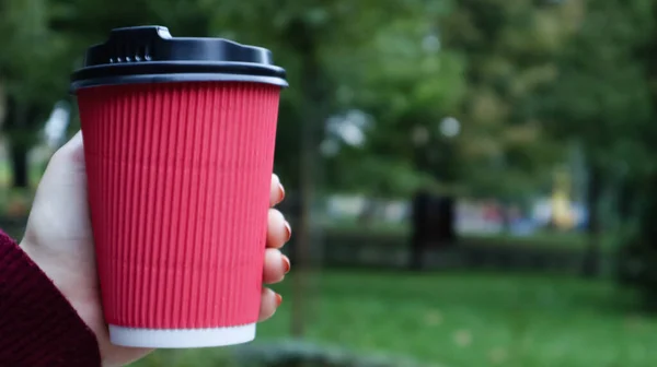 A young girl in a coat holds morning coffee with her while walking in the park. Hand holding paper cup of coffee in green park. Takeaway cappuccino. Close-up, copy space