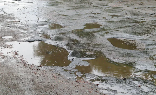 A water-filled hole in an asphalt road. A rainy day in a big city, cars are driving along the old road. Bad road with asphalt in pits and potholes that are dangerous for motorists and pedestrians