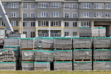 İnşaat alanında plastikle kaplı paletlerde birçok yeni gri asfalt var. Şehir caddesindeki yaya yollarından oluşan bir bina. Ön planda. Şehir meydanındaki kaldırımın tamiri