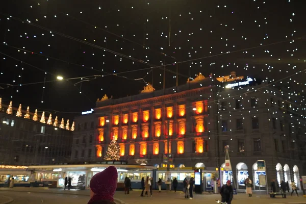 Paradeplatz 'da gece vakti kırmızı örülmüş şapka. Zürih 'teki bankaların karargahının ve tramvay istasyonunun bulanık pencereleri ve Lucy adında bir sürü asılı lamba. Zürih, 19 yaşında. Aralık 2020.