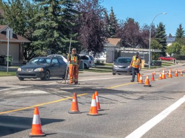 Calgary, Alberta, Kanada. 17 Ekim 2025. Güvenlik teçhizatında iki inşaat işçisi trafik konileriyle yol çalışması yapıyor. Güneşli bir caddede arabalar ve evlerle dolu..