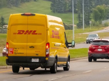 Calgary, Alberta, Kanada. 15 Ekim 2025. Bir DHL dağıtım aracı, kendine özgü sarı üniformasıyla, dünya çapında lojistik, e-ticaret ve verimli paket teslimatını sembolize eden bir yolda ilerliyor..