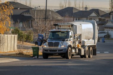 Calgary, Alberta, Kanada. 2 numara, 2025. Çöp kamyonu, banliyöde evleri ve yapraksız ağaçları olan mavi bir geri dönüşüm kutusunu kaldırmak için mekanik kol kullanıyor.