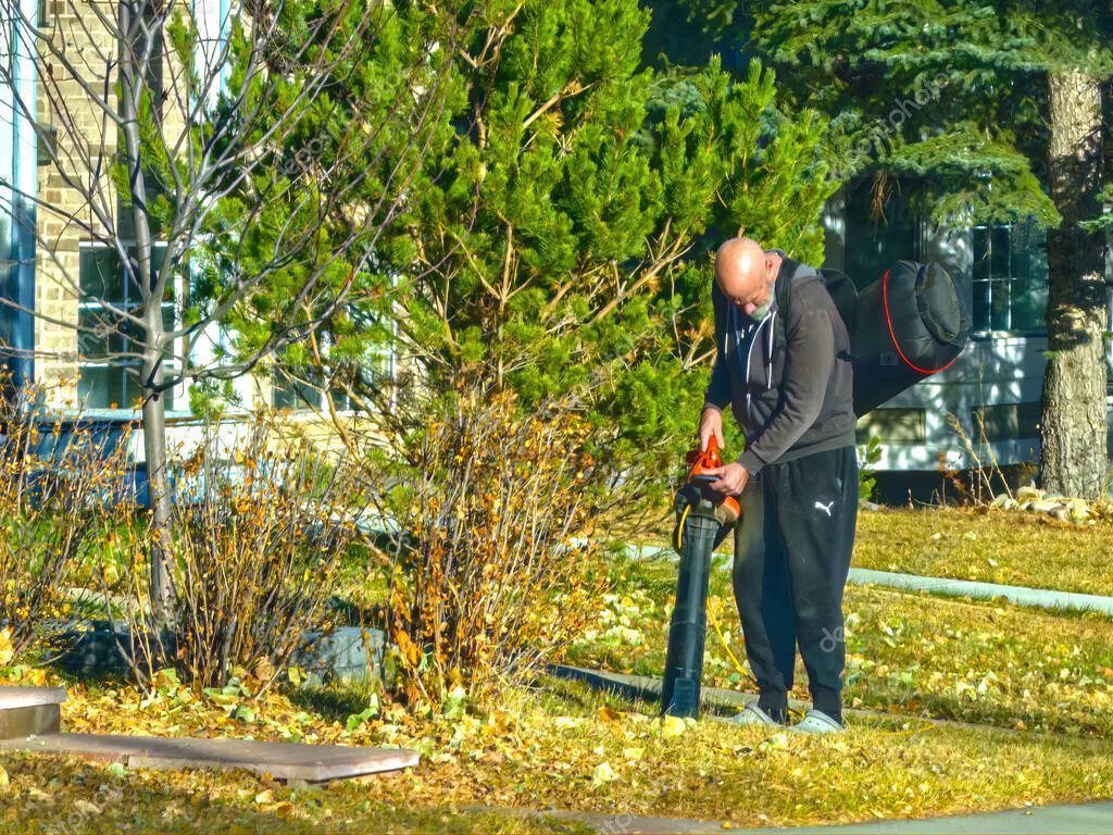 Calgary, Alberta, Canada. Nov 2, 2025. Person in Puma tracksuit uses a red-and-black leaf blower to clear autumn leaves from a residential yard