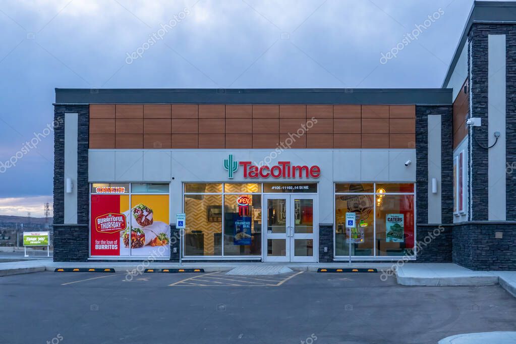 Calgary, Alberta, Canada. Nov 1, 2025. TacoTime exterior showing logo, wood and stone facade, window posters, interior seating and accessible parking under an overcast sky