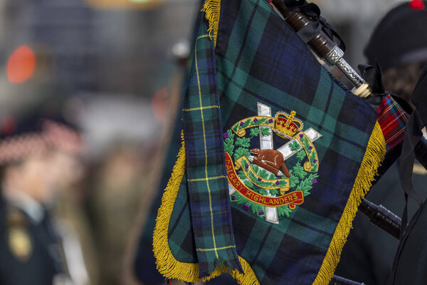 Калгари, Альберта, Канада. 11 ноября 2025 года. Крупный план канадских горцев тартан баннер с вышитым короной, бобром и поясом эмблемы, волынки и мундиры позади.