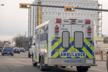 Calgary, Alberta, Kanada. 22 Kasım 2025. Arka planda büyük bir inşaat sahası olan yeşil bir trafik ışığının altından geçen, kentsel bir caddede giden bir MEDAVE ambulansının arka görüntüsü.
