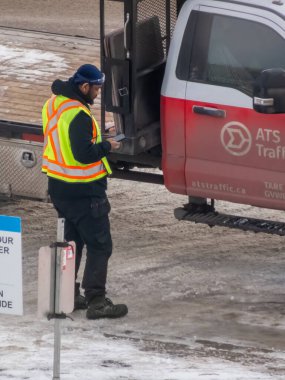 Calgary, Alberta, Kanada. 12 Aralık 2025. Yansıtıcı yelek giyen bir inşaat işçisi ATS trafik işaretli bir servis kamyonunun yanında duruyor, kışın açık hava şantiyesinde ekipmanları kontrol ediyor..
