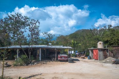 Huatulco, Oaxaca, Meksika. Aralık 18, 2025. Geniş, göz hizasında bir dış çekim, toprak bahçesi, park etmiş kırmızı arabası ve yemyeşil bir tropikal ormana karşı su deposu olan mütevazı bir evi gösteriyor..