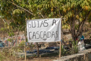 Orta seviye bir çekimde, Meksika, Oaxaca 'da yemyeşil ağaçlar ve yeşillikler arasında yerel şelale rehberlerini destekleyen ahşap kazıklara bağlanmış derme çatma bir afiş görülüyor..