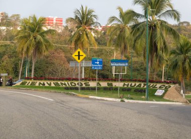 Huatulco, Oaxaca, Meksika. 14 Aralık 2025. Düşük karşılama işareti (BIENVENIDOS A B. HUATULCO) medyanın bitkilerinde, palmiye ağaçları ve yerel trafik işaretleriyle çevrili olarak hecelenir..