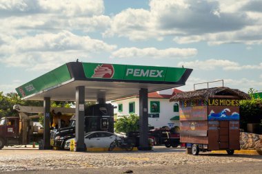 Huatulco, Oaxaca, Meksika. 17 Aralık 2025. Gündüz çekimleri yeşil bir Pemex benzin istasyonunu kaldırım taşlı bir otoparkta sazdan yapılmış bir sokak yemeği arabasının yanında gösteriyor..