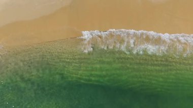 Yüksek açılı insansız hava aracı beyaz köpüklü bir okyanus dalgasının temiz, altın sarısı kumlu bir sahile vurduğu anı yakalar..