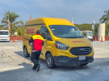 Huatulco, Oaxaca, Mexico. Dec 28, 2025. A delivery driver in uniform carries a parcel from a yellow DHL van parked in a sunny lot with palm trees and flags in the background.