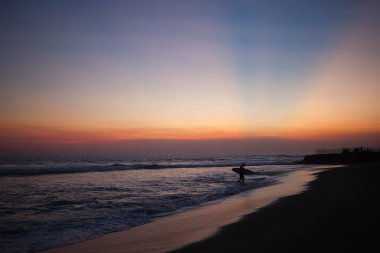 Gün batımında okyanustan çıkan bir sörfçünün silüeti. Turuncu ışınlar, Canggu, Bali, Endonezya 'daki sörf noktasının üstündeki mavi gökyüzüne nüfuz ediyor..