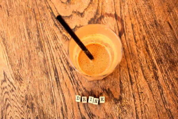 Latte coffee with ice and straw in a glass on a wooden table and the word 'drink' written with wooden cubicles, top view, flat lay.