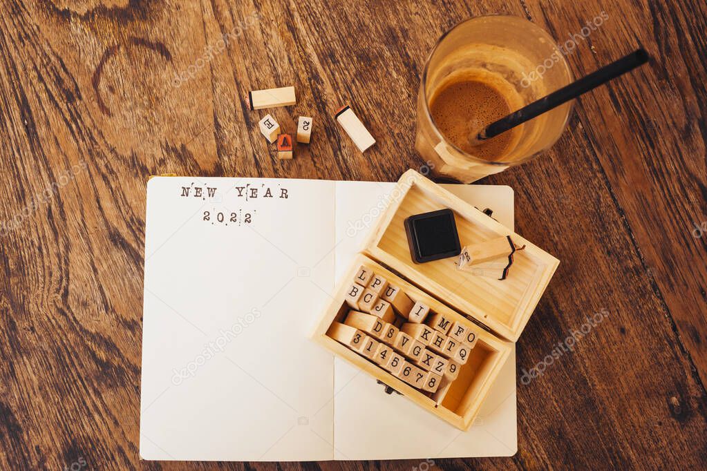New Year 2022 printed on a diary page with a stamp letters set and ink, coffee latte and scattered stamps on a wooden desk in a cafe. Scrapbooking for new year resolutions, top view, flat lay.