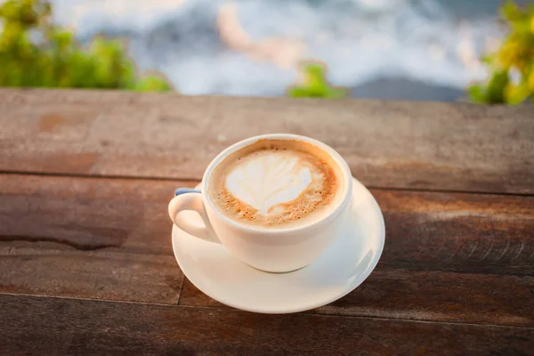 Delicious cup of coffee latte on a wooden table in a restaurant with ocean view. Perfect hot drink for a holiday-maker.