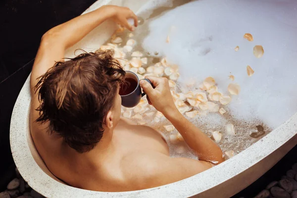 Homem deitado na banheira com espuma de sabão, pétalas, caneca — Fotografia de Stock